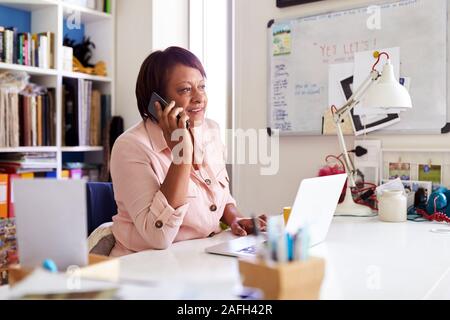 Mature Woman With Laptop Working In Home Office Using Mobile Phone Stock Photo