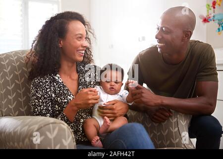 Loving Parents Sitting In Chair Cuddling Baby Son In Nursery At Home Stock Photo