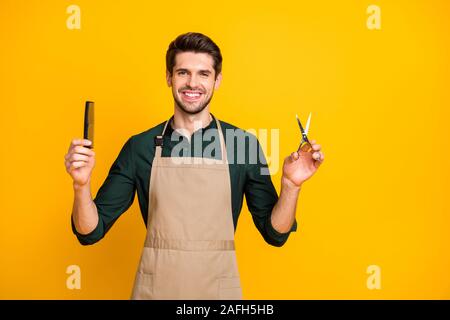 Portrait of positive cheerful guy freelancer ready to make haircut to his clients in modern barbershop hold comb scissors wear stylish trendy green Stock Photo
