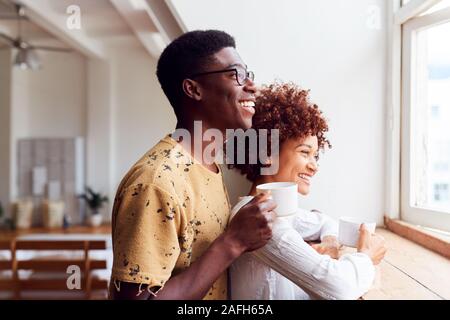 Young Couple Relaxing In Loft Apartment Looking Out Of Window With Hot Drink Stock Photo