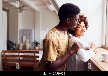 Young Couple Relaxing In Loft Apartment Looking Out Of Window With Hot Drink