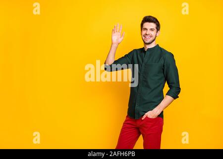 Photo of cheerful positive brown haired confident guy holding hand in pocket of red trousers smiling toothily waving his hand at you isolated vibrant Stock Photo
