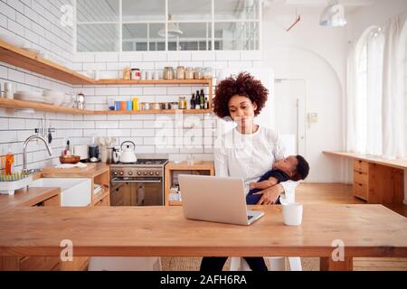 Multi-Tasking Mother Holds Sleeping Baby Son And Works On Laptop Computer In Kitchen Stock Photo