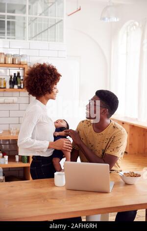 Busy Family In Kitchen At Breakfast With Father Caring For Baby Son Stock Photo