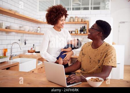 Busy Family In Kitchen At Breakfast With Father Caring For Baby Son Stock Photo