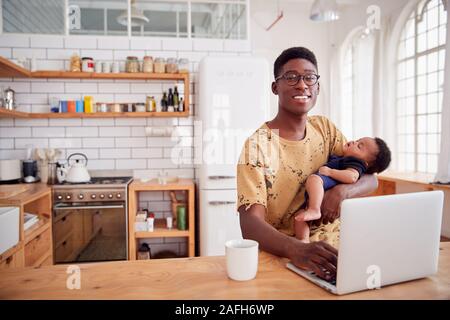 Portrait Of Multi-Tasking Father Holding Sleeping Baby Son And Working On Laptop Computer In Kitchen Stock Photo