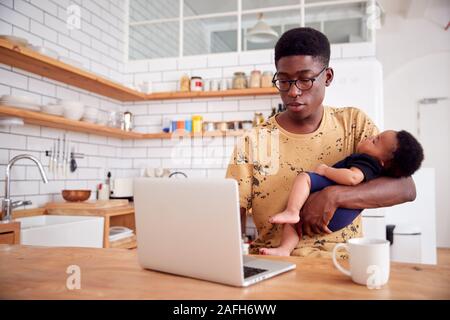 Multi-Tasking Father Holds Sleeping Baby Son And Works On Laptop Computer In Kitchen Stock Photo