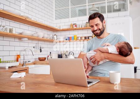 Multi-Tasking Father Holds Sleeping Baby Son And Works On Laptop Computer In Kitchen Stock Photo