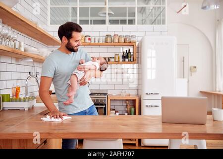 Multi-Tasking Father Holds Sleeping Baby Son And Cleans In Kitchen Stock Photo