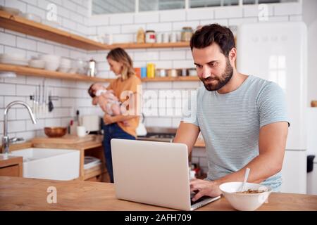 Multi-Tasking Mother Holds Baby Son And Makes Hot Drink As Father Uses Laptop And Eats Breakfast Stock Photo