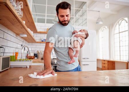 Multi-Tasking Father Holds Sleeping Baby Son And Cleans In Kitchen Stock Photo