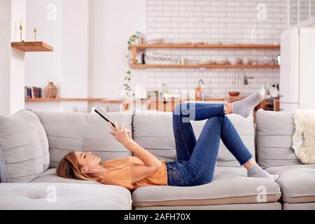 Woman Relaxing Lying On Sofa At Home Looking At Digital Tablet Stock Photo