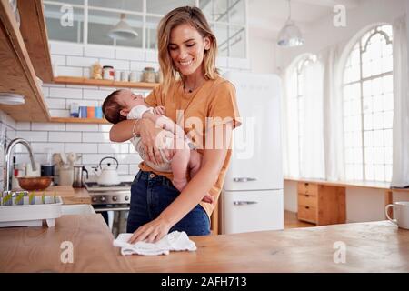 Multi-Tasking Mother Holds Sleeping Baby Son And Cleans In Kitchen Stock Photo