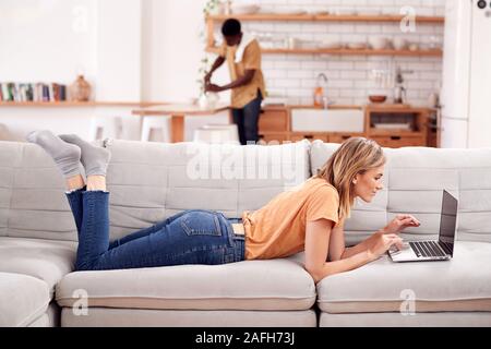 Woman Relaxing Lying On Sofa At Home Using Laptop Computer With Man In Kitchen Behind Stock Photo
