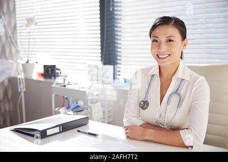 Portrait Of Smiling Female Doctor With Stethoscope Sitting Behind Desk In Office Stock Photo