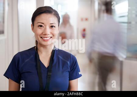 Portrait Of Smiling Female Nurse Wearing Scrubs In Busy Hospital Corridor Stock Photo