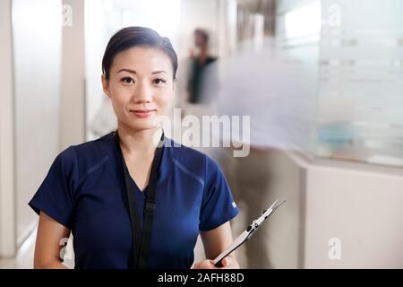 Portrait Of Female Nurse Wearing Scrubs With Clipboard In Busy Hospital Corridor Stock Photo