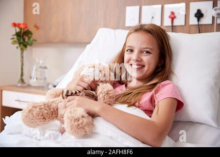 Portrait Of Girl Lying In Bed In Hospital Ward Hugging Teddy Bear Stock Photo