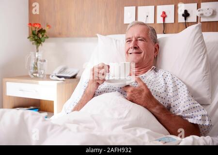 Male Senior Patient Lying In Hospital Bed Enjoying Hot Drink Stock Photo