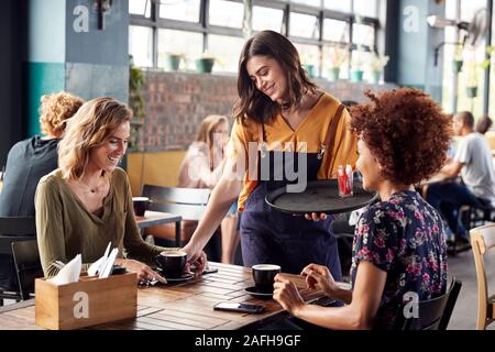 Two Female Friends Sitting At Table In Coffee Shop Being Served By Waitress Stock Photo
