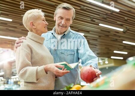 Waist up portrait of smiling senior couple choosing fresh fruits and vegetables while enjoying grocery shopping in supermarket, copy space Stock Photo