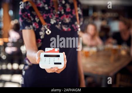 Close Up Of Waitress Holding Credit Card Payment Terminal In Busy Bar Restaurant Stock Photo