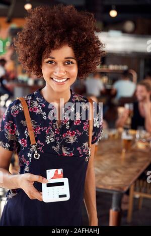 Portrait Of Waitress Holding Credit Card Payment Terminal In Busy Bar Restaurant