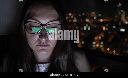 Portrait of young brunette woman in glasses working on computer near the window with cityscape view. Overtime work at night, young careerist. Stock Photo