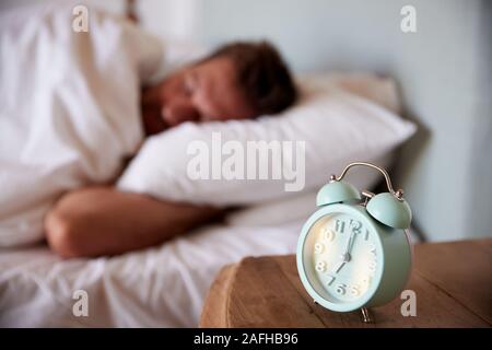 Mid adult man asleep in bed, alarm clock on the bedside table in the foreground, focus on foreground Stock Photo