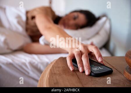 Mid adult woman lying in bed, reaching out to smartphone on the bedside table in the foreground Stock Photo