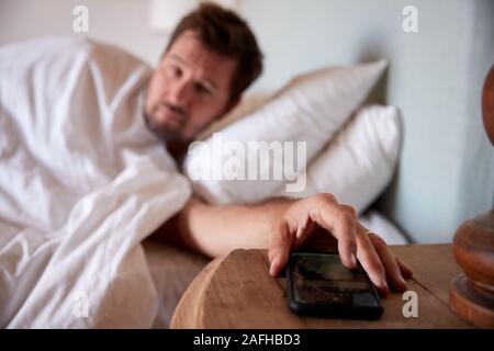 Mid adult man lying in bed, reaching out to smartphone on the bedside table in the foreground Stock Photo