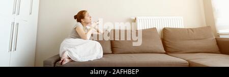 Young brunette woman in her modern apartment in living room. She turns on the air conditioner from the remote control sitting on the sofa. Climate control at home with split system, toned Stock Photo
