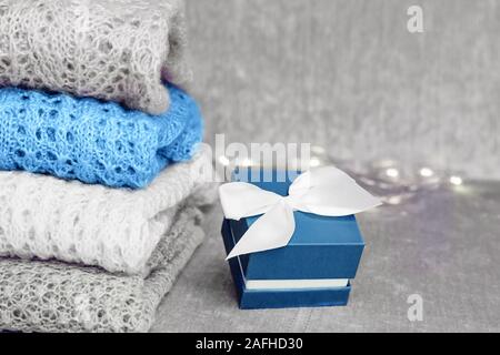 Stylish knitted pastel colored sweaters folded in stack and trendy blue colored present box with white satin bow on velvety fabric background. Winter Stock Photo