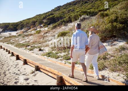 Senior white couple walking along wooden promenade on a beach holding hands, full length, back view Stock Photo