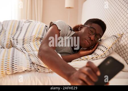 Millennial African American man wearing glasses, half asleep in bed holding smartphone, close up Stock Photo