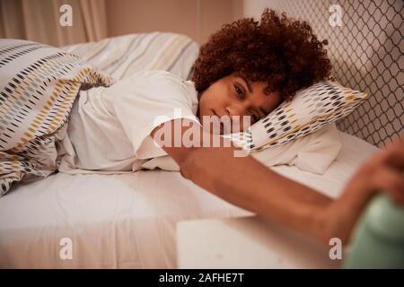 Millennial African American woman half asleep in bed, reaching out to stop alarm clock, close up