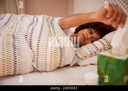Millennial African American woman lying ill in bed, taking a tissue from a box, side view, close up