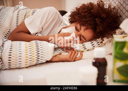 Millennial African American woman lying in bed blowing her nose into a tissue, side view, close up