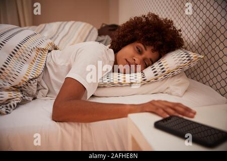 Millennial African American woman half asleep in bed, reaching out for her smartphone, close up