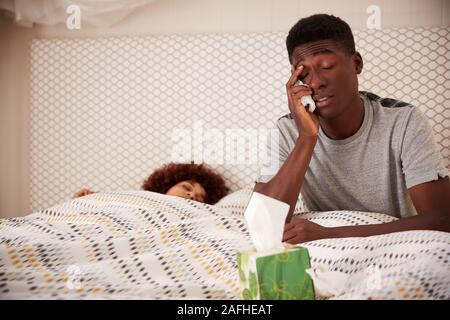 Millennial African American man ill in bed, sitting up while partner sleeps beside him, close up Stock Photo