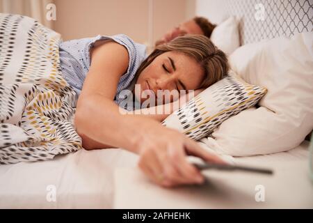 White woman half asleep in bed holding smartphone, her partner sleeping in background, close up Stock Photo