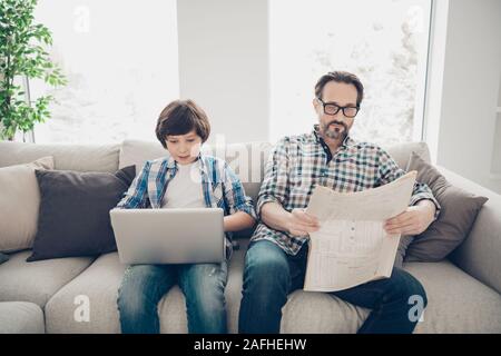 Portrait of two nice attractive lovely cute focused concentrated guys dad and pre-teen son sitting on sofa using laptop reading news in light white Stock Photo