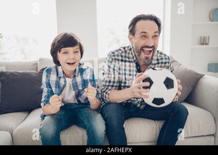 Portrait of two nice attractive lovely cute cheerful cheery overjoyed guys dad and pre-teen son sitting on sofa watching sport show online in light Stock Photo