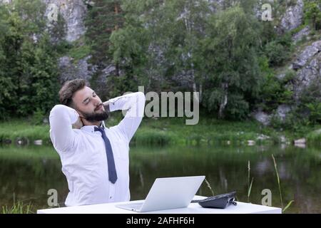 young male office worker massaging his neck while sitting at a workplace. Businessman feeling pain in neck after sitting at desk. Feeling exhausted. Stock Photo