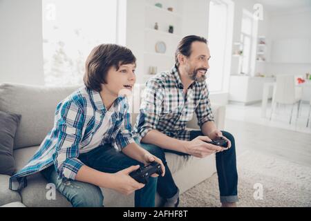 Profile side view portrait of two nice attractive cheerful cheery guys dad and pre-teen son sitting on sofa playing online game spending weekend at Stock Photo