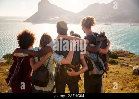 Millennial friends on a hiking trip reach the summit and embrace, admiring the view, back view Stock Photo