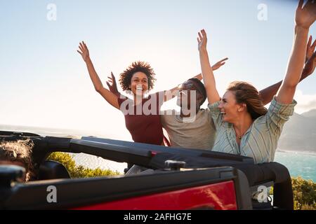 Excited millennial friends travelling in the back of an open car with their arms in the air Stock Photo