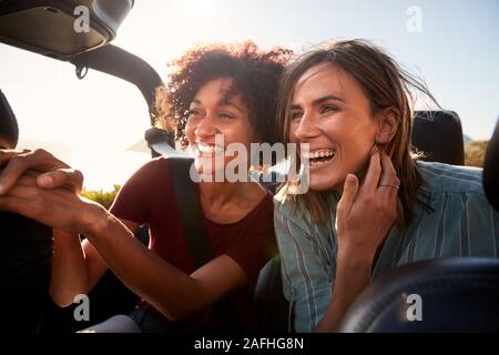 Two millennial female friends on a road trip smiling in the back of an open top car, close up Stock Photo