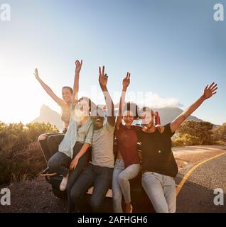 Five millennial friends on a road trip taking a break, leaning on the car waving to camera Stock Photo
