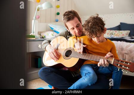 Single Father At Home With Son Teaching Him To Play Acoustic Guitar In Bedroom Stock Photo